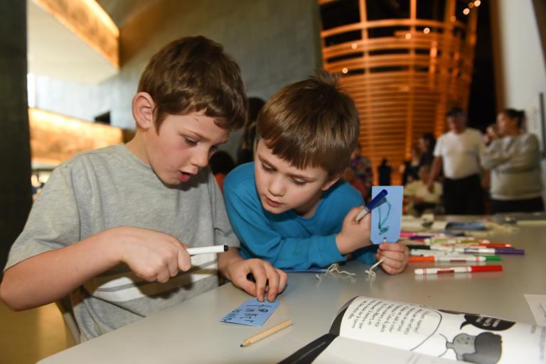 Two young boys holding markers and small cards with string and reading one of the cards. Visibilité masquée.