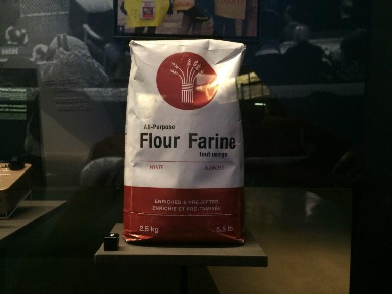 Un sac de farine dans une exposition. Le texte sur le sac lit comme suit : All-Purpose Flour/Farine tout usage, White/Blanche, Enriched & Pre-Sifted/Enrichie et pré-tamisée, 2.5 kg/5.5lb