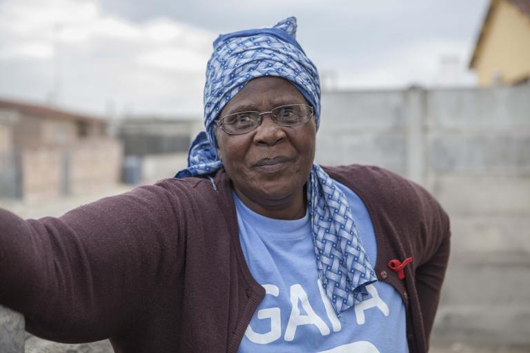 A head-and-shoulders portrait of Gogo Gladys Tyophol. She is wearing glasses and a blue patterned kerchief wrapped around her head. Her shirt says GAPA and a red AIDS ribbon is attached to her sweater. Visibilité masquée.