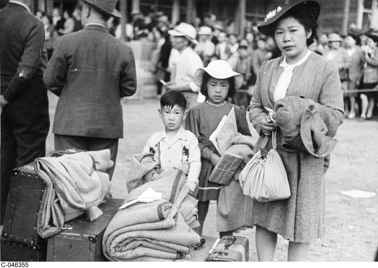 Photo en noir et blanc d’une femme avec deux enfants qui se tiennent derrière une pile de bagages et de couvertures et qui fixent l’objectif. Visibilité masquée.