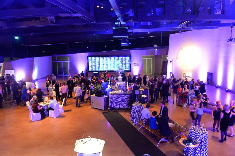 Une fête se déroule dans une grande salle. Les gens sont rassemblés autour de tables et se parlent entre eux en tenant des verres. La salle est éclairée de lumières violettes et bleues. Visibilité masquée.