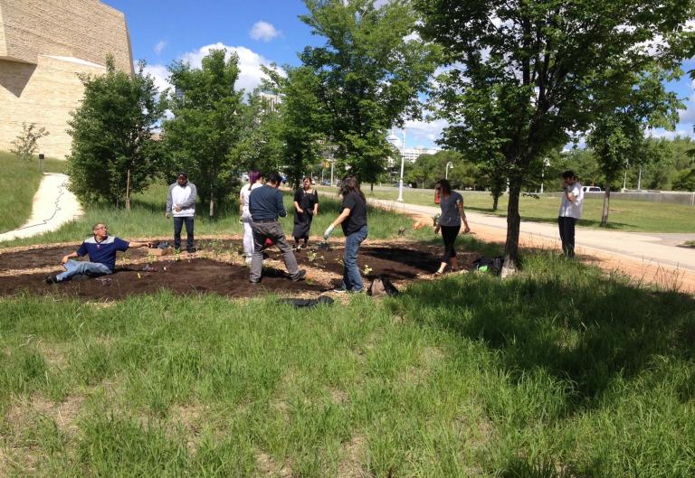 Sept personnes, hommes et femmes, travaillent dans un jardin par une journée ensoleillée. Le jardin est circulaire et renferme bien peu de plantes. Autour du jardin, on voit des herbes hautes, des arbres, un sentier et un trottoir. Visibilité masquée.