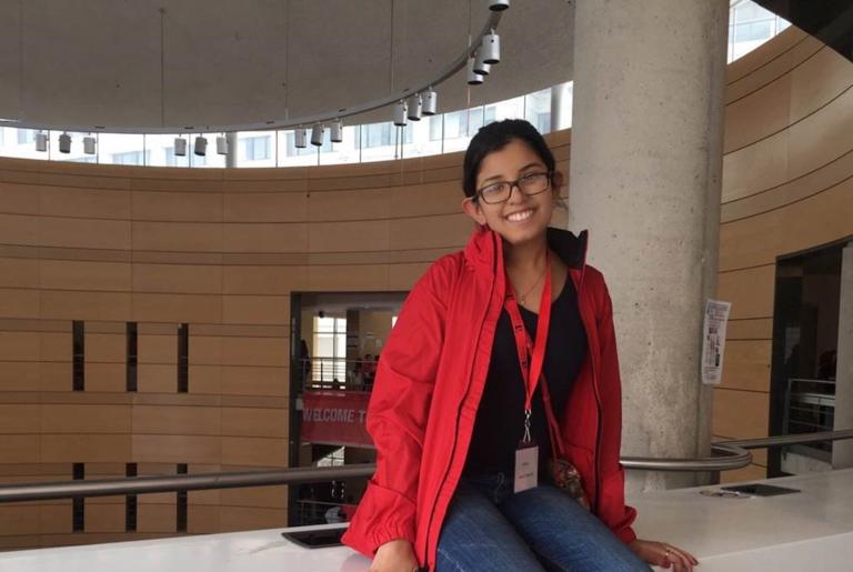 Une jeune femme est assise sur un rebord dans une grande salle circulaire. Elle sourit au photographe et porte un jeans, une chemise foncée et un manteau rouge. Visibilité masquée.