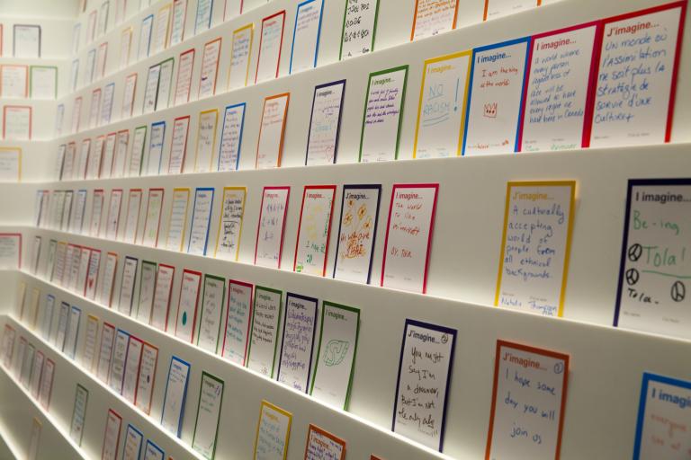 Des cartes avec l’expression « J’imagine… » en français et en anglais sont placées le long d’un mur blanc sur plusieurs rangées de présentoirs. Des personnes en visite au Musée ont dessiné ou écrit sur chaque carte avec des marqueurs. Visibilité masquée.