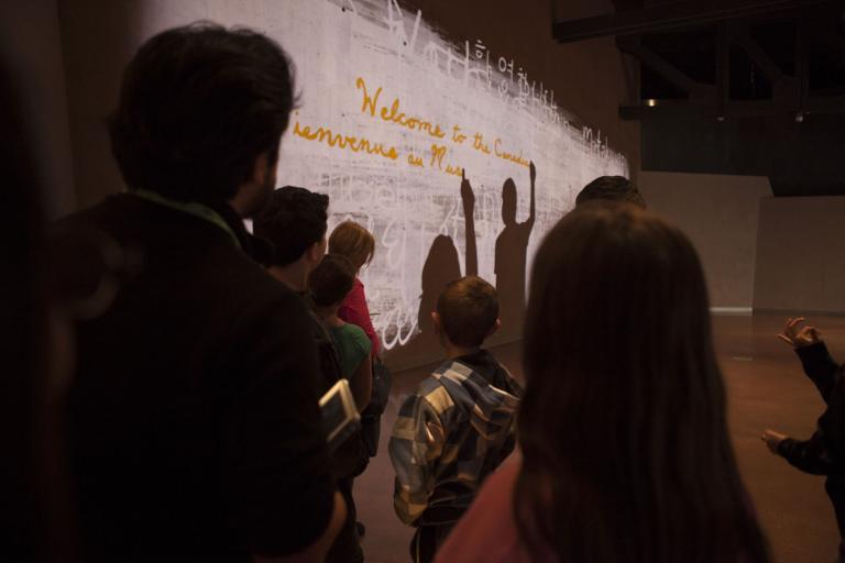 Un groupe de personnes devant un mur avec une projection de deux silhouettes en train d'écrire la phrase « Bienvenue au Musée canadien pour les droits de la personne » en anglais et en français. Visibilité masquée.