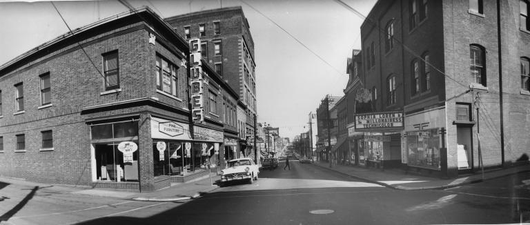 Une image en noir et blanc d’une rue de ville. La rue s’éloigne de nous vers un point invisible à l’horizon. De chaque côté de la rue se trouvent des bâtiments en brique de deux à sept étages de hauteur. Beaucoup des bâtiments ont des vitrines au rez-de-chaussée et le premier à droite a un baldaquin de cinéma qui annonce le film « The Millionairess », mettant en vedette Sophia Loren.