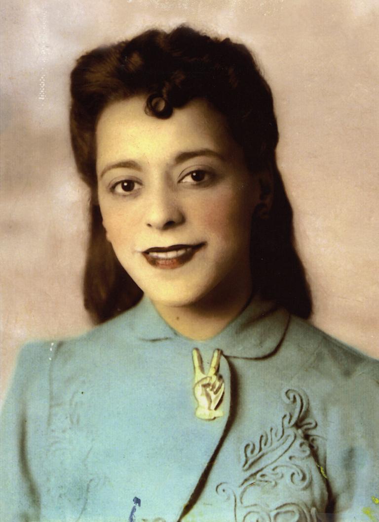Portrait en buste d’une Viola Desmond souriante. Elle porte une veste bleu pâle à motifs brodés. La veste est fermée au col par une broche en forme de main avec deux doigts écartés pour former le V de la victoire. Visibilité masquée.