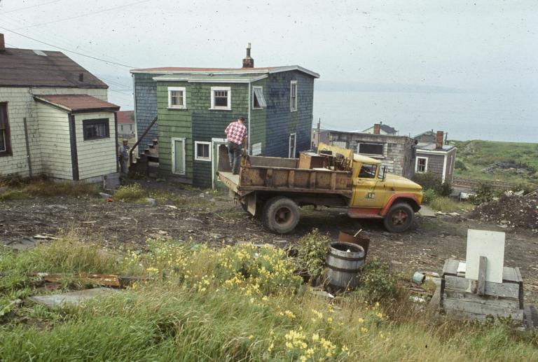 Un homme se tient à l’arrière d’un camion à ordures garé devant une maison en bois aux bardeaux verts. À l’arrière-plan, on aperçoit des rails de chemin de fer et d’autres maisons, et plus loin encore, le port d’Halifax.