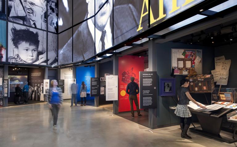 Une personne se promène dans une galerie du Musée, tandis que six autres se tiennent dans ou près des alcôves qui bordent le mur de la galerie. Chaque alcôve contient une exposition différente avec du texte ou des images. Visibilité masquée.