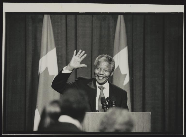 Une photographie en noir et blanc de Nelson Mandela debout derrière un podium. Il sourit et fait signe de la main. Visibilité masquée.