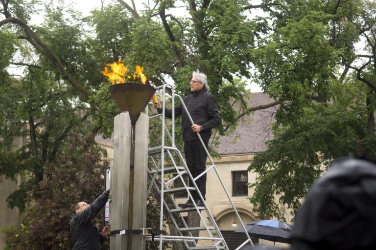 Un homme tenant un flambeau est debout sur une échelle pour allumer une flamme dans une vasque en cuivre. Visibilité masquée.