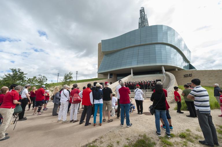 Un groupe de personnes regardant une chorale qui chante sur les marches de l’amphithéâtre extérieur du Musée. Visibilité masquée.