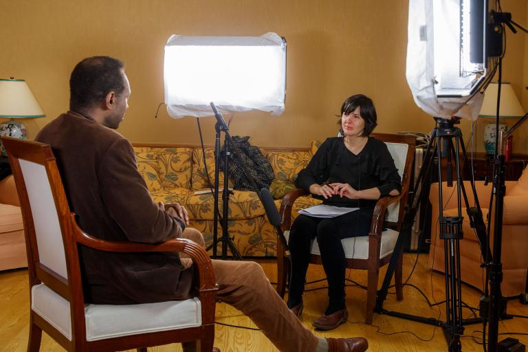 Un homme et une femme sont assis face à face dans des chaises. On voit un microphone sur pied entre eux, ainsi que de grandes lampes de chaque côté de la chaise de la femme. Les lampes sont pointées vers l’homme. Visibilité masquée.