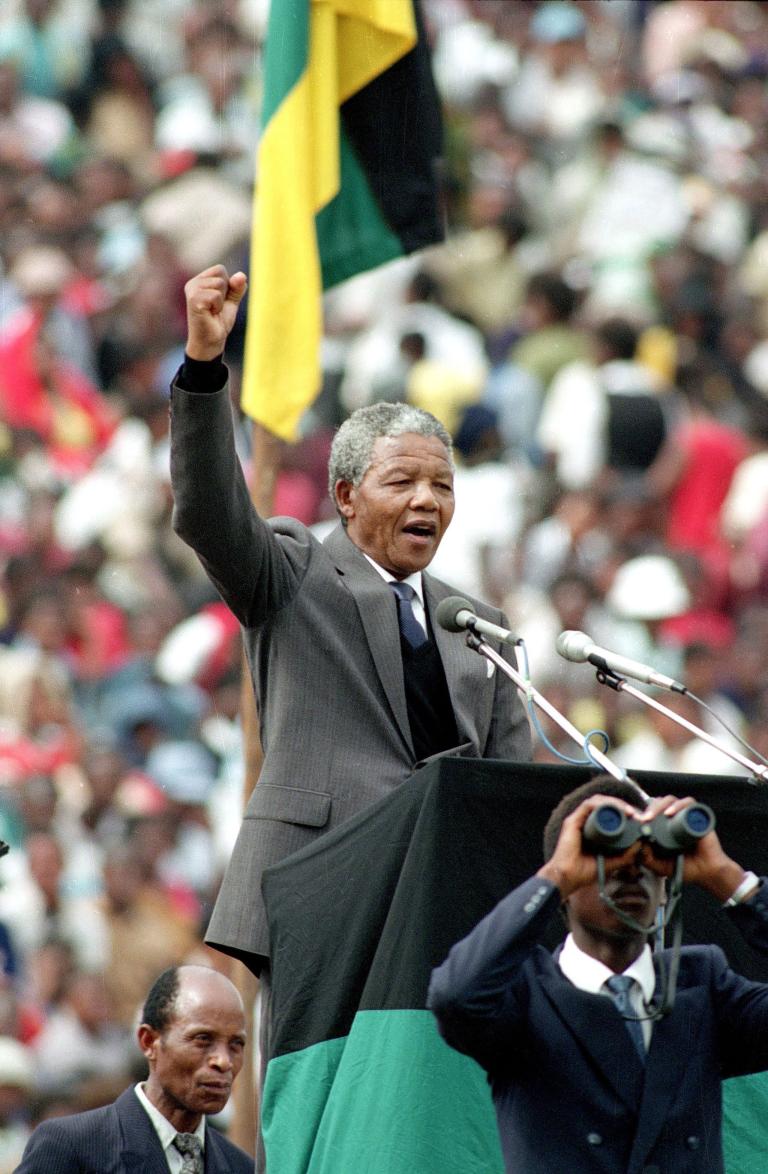 Nelson Mandela, un homme plus âgé à la peau foncée et aux cheveux gris et courts, est debout sur un podium et parle dans deux microphones. Il porte un complet gris et une cravate. Il lève son poing droit au-dessus de sa tête. Visibilité masquée.