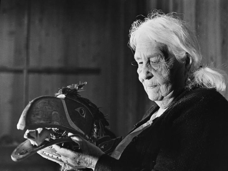 Une image en noir et blanc d’une femme âgée aux cheveux blancs tenant un masque formé et peint de façon à représenter la tête d’un oiseau. Le long bec est ouvert, et il y a des plumes attachées sur le haut du masque. La femme regarde le masque qu’elle tient dans ses mains. Visibilité masquée.