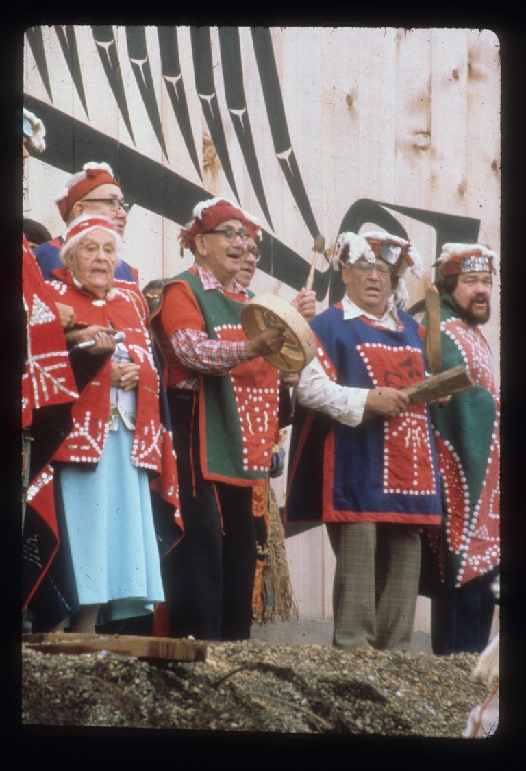 Un groupe d’hommes et de femmes plus âgés chante, debout devant un grand bâtiment en bois. Un homme joue du tambour. Tous et toutes portent de longues chemises ou des couvertures décorées de motifs faits de boutons.