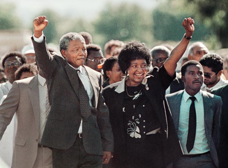Nelson Mandela, un homme noir âgé aux cheveux grisonnants, forme un poing levé avec sa main droite et le tient au-dessus de sa tête. De sa main gauche, il tient la main de Winnie Mandela, une femme noire aux cheveux noirs. Elle sourit et forme un poing levé avec sa main gauche, à l'image du poing levé de Nelson Mandela. Une foule nombreuse est rassemblée derrière eux. Visibilité masquée.