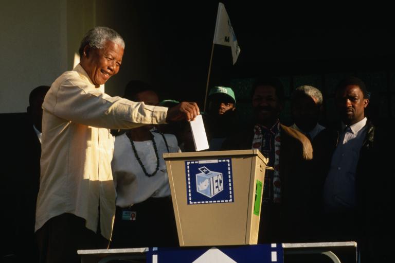 Un Nelson Mandela âgé sourit en tendant le bras droit pour déposer son bulletin de vote dans une grande boîte de scrutin en métal.