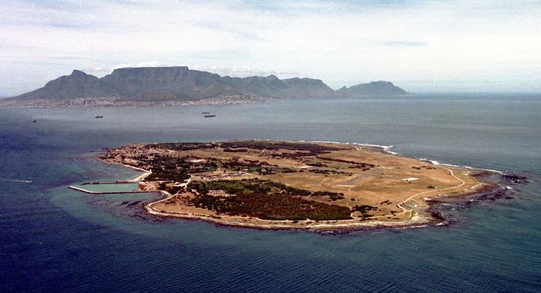 Vue aérienne de l’île Robben. L’île est plus ou moins de forme ovale et le terrain est très plat et couvert presqu’à moitié d’arbres. On aperçoit un terrain d’aviation sur le côté droit de l’île. Au loin, derrière l’île, on voit le continent, y compris les édifices d’une ville. Au-delà de la ville se dressent de grandes montagnes.