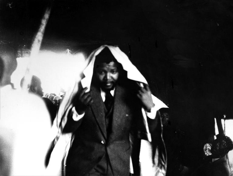 Une image en noir et blanc d’un jeune Nelson Mandela portant un complet, voûtant le dos et tenant un imperméable autour de sa tête, comme pour se protéger de la pluie.