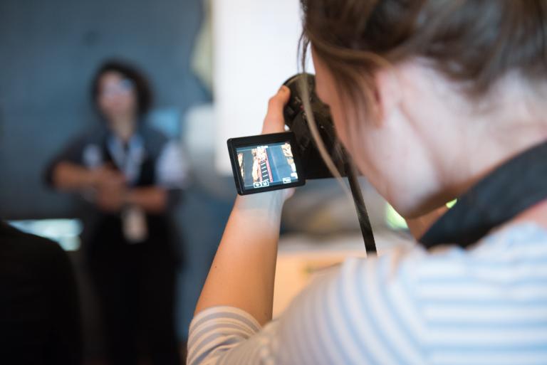 A person looks through the screen of a camera in the foreground while a person poses in the background. Visibilité masquée.