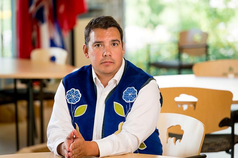 A man wearing a blue vest with flower beadwork, seated with his hands resting on a table. Visibilité masquée.