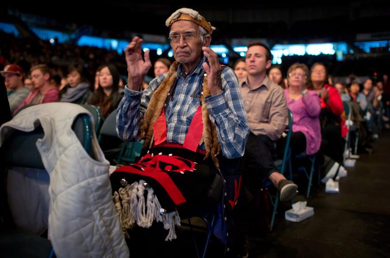 Un homme âgé vêtu d'une chemise à carreaux et d'accessoires autochtones traditionnels est assis dans une foule. Il a les bras pliés aux coudes et les mains levées, ouvertes l’une face à l’autre.