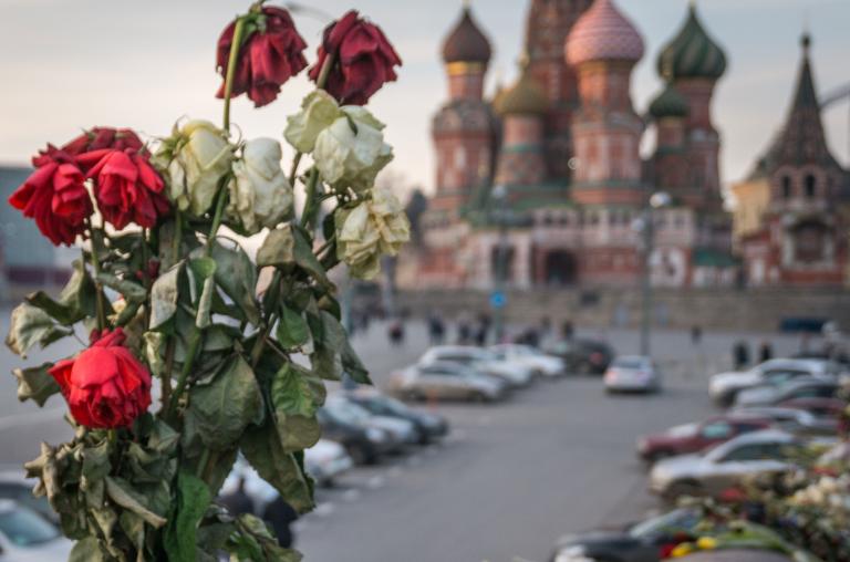 Des roses rouges et blanches fanées dans un vase. En arrière-plan, on voit la cathédrale Saint-Basile sur la place Rouge, à Moscou, avec ses multiples coupoles et dômes, qui rappellent un feu de camp montant vers le ciel. Visibilité masquée.