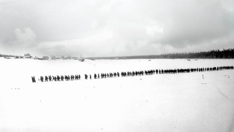 Une photo en noir et blanc d’un paysage plat et enneigé. On voit des arbres et des petits bâtiments au loin. Une longue file de plus de 100 personnes marchent dans la neige. Visibilité masquée.
