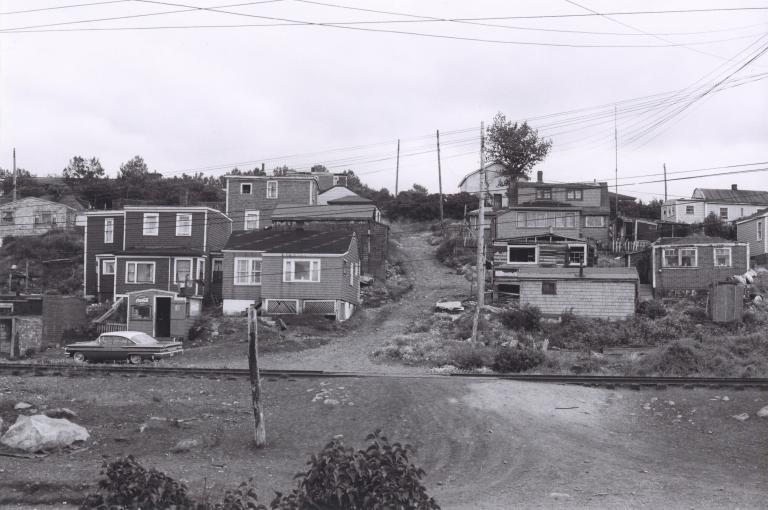 Une image en noir et blanc d’une douzaine de maisons sur une colline. Au milieu de l’image, on voit un chemin de terre qui monte la colline entre les maisons.