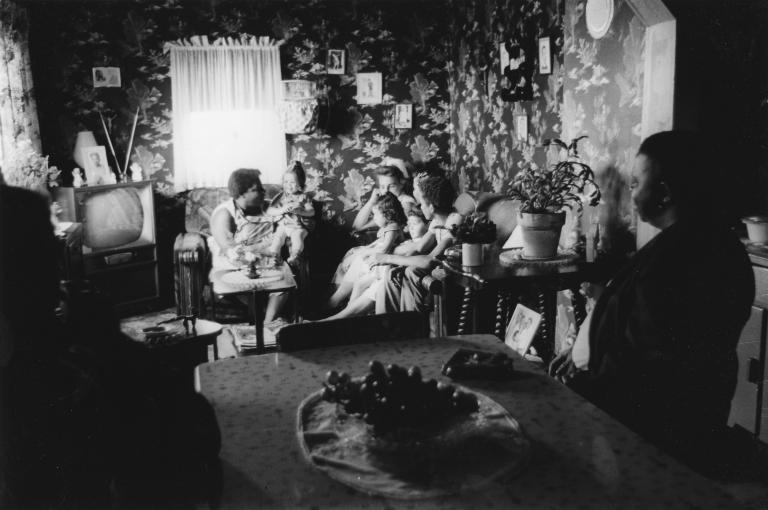 Une image en noir et blanc de femmes et de jeunes enfants assis sur des sofas et des chaises dans un petit salon.