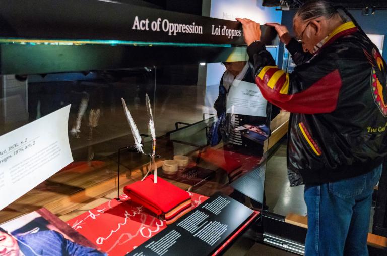 Un homme aux longs cheveux tressés et à la tête penchée se tient devant une vitrine où se trouvent deux plumes. Visibilité masquée.