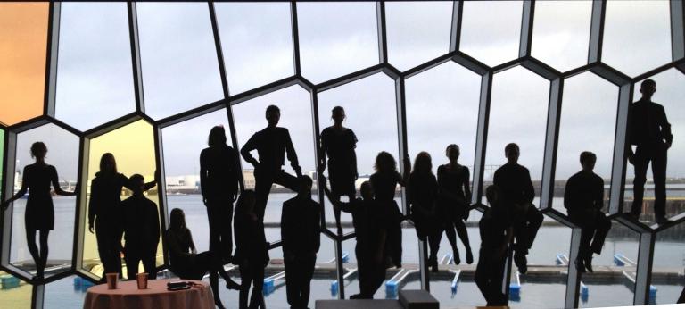 Dix-huit personnes, certaines assises, certaines debout, placées en rangée le long d’un mur de fenêtres aux formes irrégulières. Visibilité masquée.