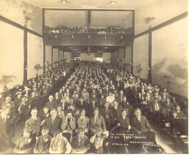 Photo en noir et blanc d’un public de cinéma. La photo est prise de l’avant du cinéma en regardant vers l’arrière, de sorte que les visages du public sont visibles. Des palmiers en pot bordent les murs de chaque côté. Visibilité masquée.