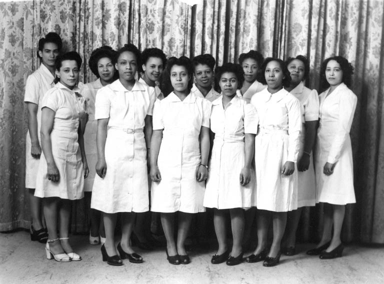 Une image en noir et blance de Viola Desmond avec 11 autres femmes. Elles portent toutes des robes d’un blanc uni et se tiennent debout en deux rangées. Elles regardent vers l’objectif. 