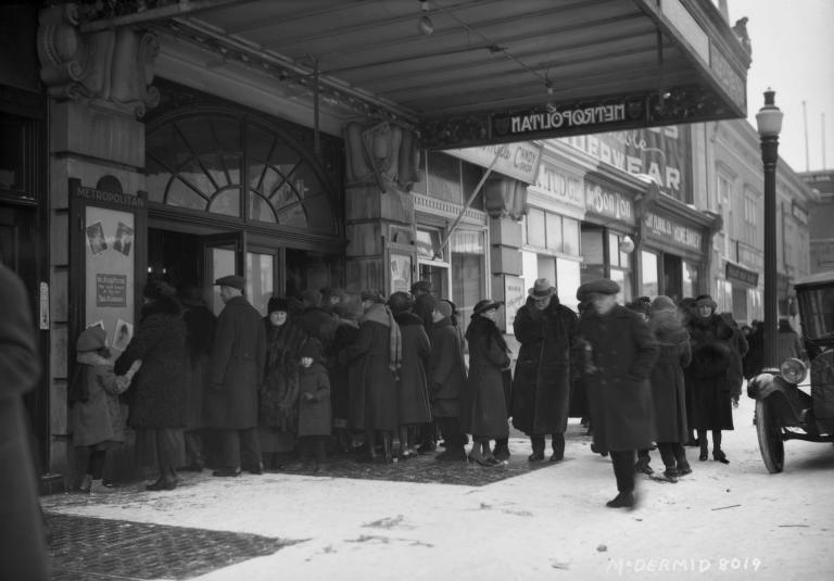 Une mage en noir et blanc d’une foule devant l’entrée d’un théâtre du nom de « Metropolitan ». Tout le monde porte de longs manteaux à l’ancienne, et certains portent des chapeaux. Une voiture antique est garée dans la rue.