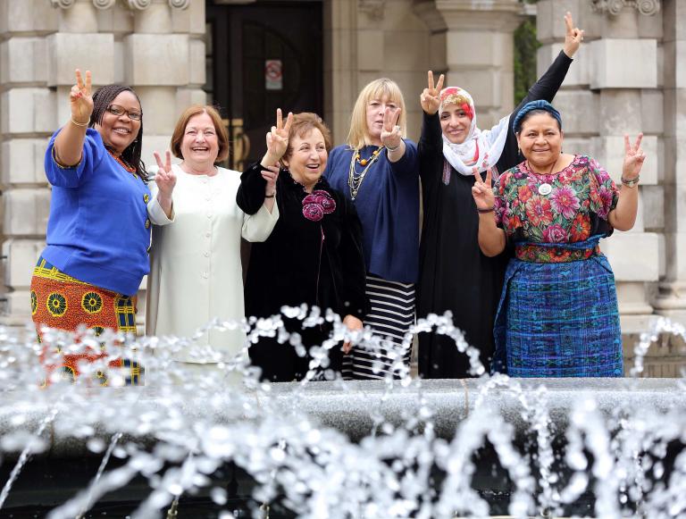 Six femmes debout devant ce qui semble être l’entrée d’un grand édifice de pierre. Elles sourient toutes et lèvent une ou deux mains en faisant le signe de la paix.