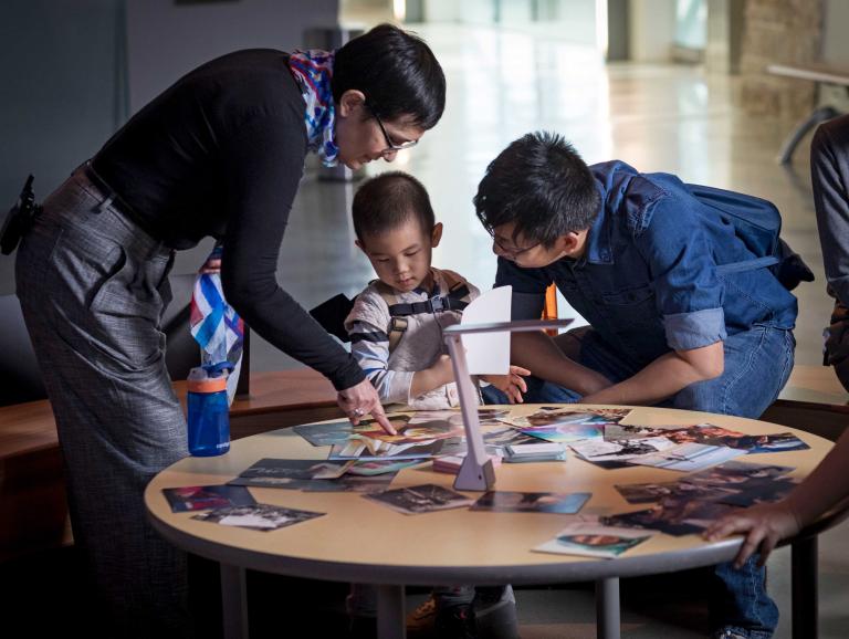 Une femme, un homme et un jeune garçon sont rassemblés autour d’une table circulaire. Plusieurs photographies sont éparpillées sur la table et la femme en montre une au garçon. Visibilité masquée.