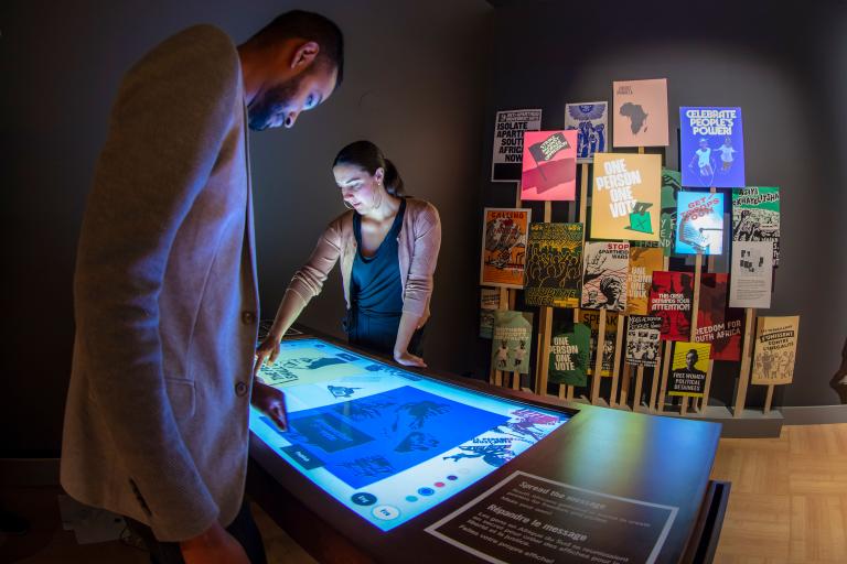 Un homme et une femme regardent une table lumineuse. À l'arrière-plan, on voit un groupe d'affiches colorées sur le mur. Visibilité masquée.