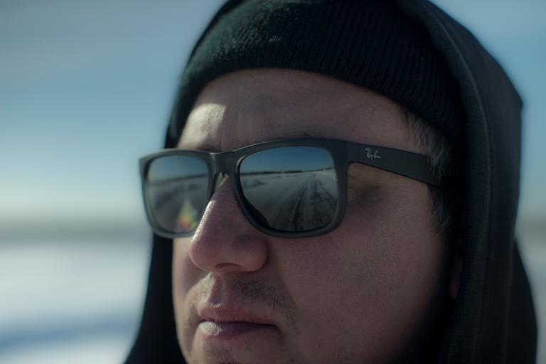 Un gros plan d'un homme portant des lunettes de soleil et un chapeau, qui regarde vers quelque chose hors champ. On voit le reflet d'une route dans ses lunettes. Visibilité masquée.