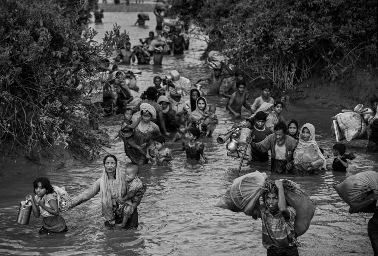 Des femmes, des enfants et des hommes rohingyas marchent dans l’eau d’une rivière qui leur monte jusqu’à la taille. Certaines personnes portent des jeunes enfants alors que d’autres portent des sacs de possessions, dont des articles ménagers. Visibilité masquée.