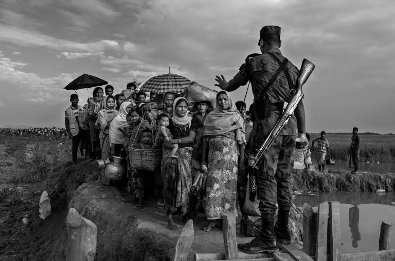Un soldat avec un fusil en bandoulière sur le dos empêche un groupe de Rohingyas avant qu’ils ne traversent un pont de fortune. Sa main gauche est levée et fait un geste pour qu'ils s'arrêtent.