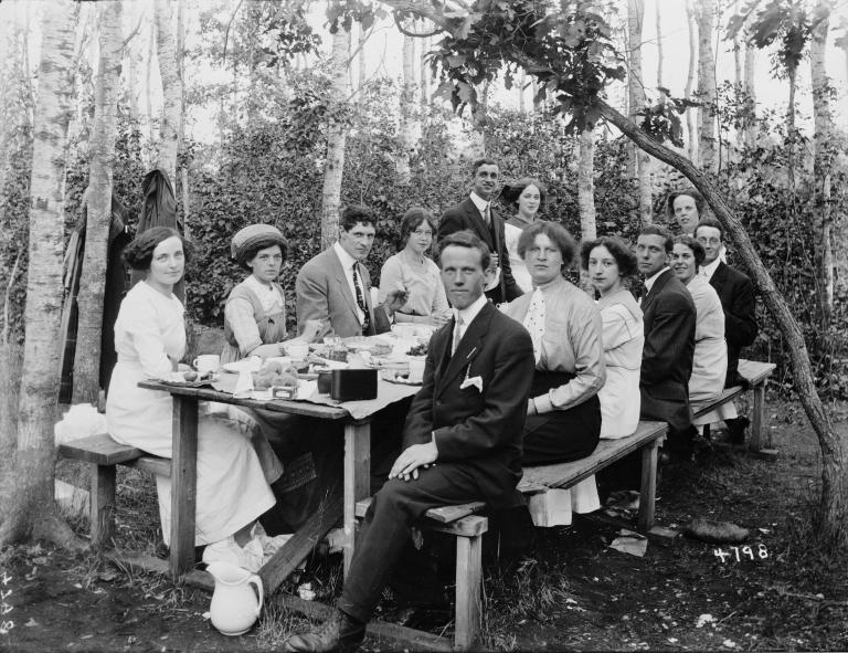 Un groupe d’hommes et de femmes bien habillés assis à une table de pique-nique avec des arbres en arrière-plan. 