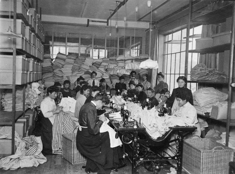 Un grand groupe de femmes assises en train de coudre. D’autres sont debout derrière elles dans une salle remplie de tissus.
