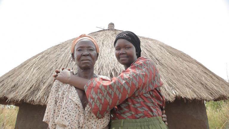 Deux femmes debout devant une hutte avec un toit de paille, regardent la caméra. La femme plus à droite sourit en serrant l’autre femme dans ses bras.