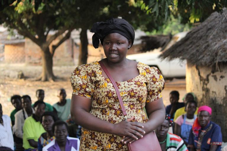 Une femme portant un haut à fleurs brun et jaune est représentée à partir de la taille, avec plusieurs personnes assises derrière elle dans un décor extérieur.