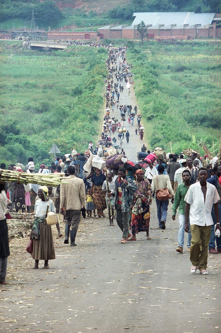 Une grande foule de personnes de tous âges portant de la nourriture et des effets personnels se dirige vers la caméra sur un long chemin de terre traversant un paysage vert vif d’herbe et de buissons. La route et la foule s’étendent dans le lointain. Visibilité masquée.