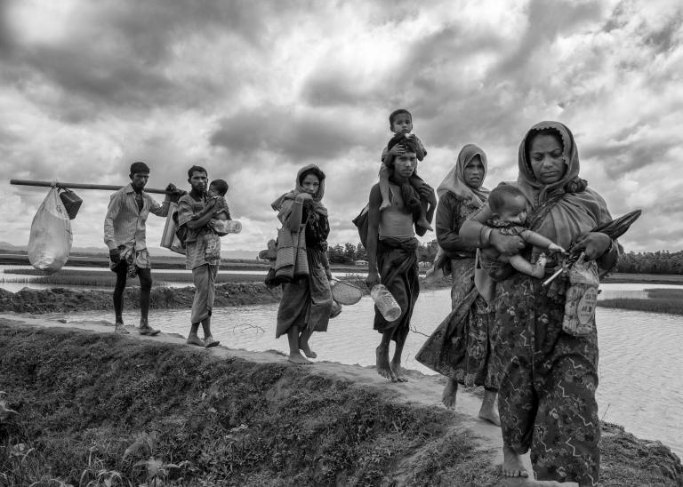 Un groupe de femmes et d’hommes rohingyas transportent de jeunes enfants et des effets personnels en marchant sur une digue de terre qui traverse une étendue d’eau. Visibilité masquée.