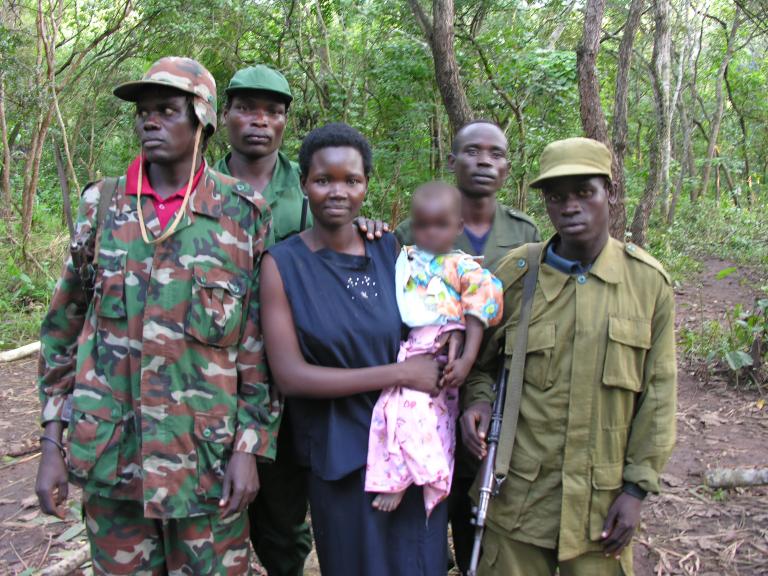 Une femme tenant un bébé, tandis que quatre hommes en tenue militaire se tiennent à côté et derrière elle. Ils sont tous debout devant une forêt et posent pour la photo. Visibilité masquée.