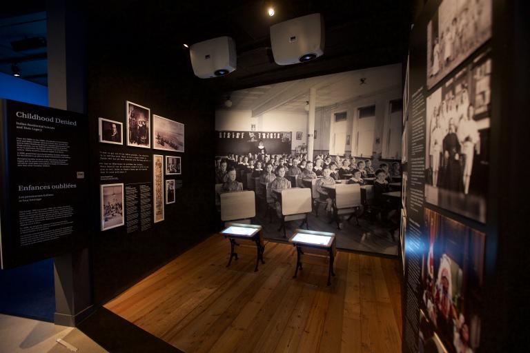 Une exposition muséale montrant une photo en noir et blanc d’enfants assis à des pupitres placés en rangées. Deux pupitres semblables à ceux qu’on voit dans la photo sont placés au centre de l’exposition. Sur un panneau de texte, on peut lire « Enfances oubliées ». Visibilité masquée.